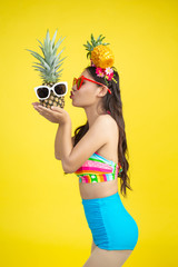 Beautiful woman in a swimsuit holding a pineapple poses on a yellow background.