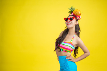Beautiful woman in a swimsuit holding a pineapple poses on a yellow background.