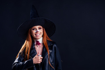 redhaired ginger girl dressed with a huge witch hat and black leather jacket ,spooky blood make up fracture of nose and neck bandage in studio background