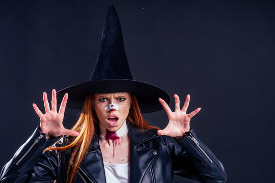 redhaired ginger woman witch black hat and in leather jacket screaming and scaring with crazy make up on her pretty face in studio pink background