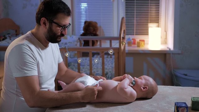 Dad changes diaper pants baby. A young caucasian father with a beard and glasses changes the diaper of his child. Medium shot