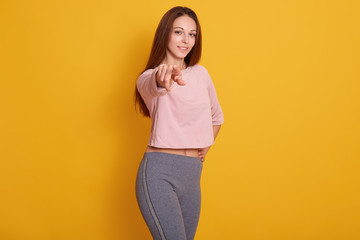 Portrait of young Caucasian athletic girl pointing at camera, looking smiling at camera, wearing stylish sporty clothing, model posing isolated over yellowstudio background.Fitness and sport concept.