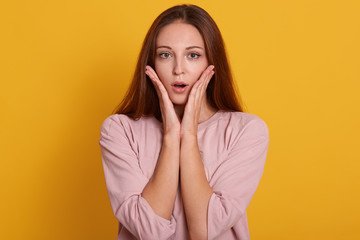 Close up portrait of emotional female being shocked to see something, opens mouth with surprisment and keepspalms near cheek, poses against yellow studio background. People emotions concept.