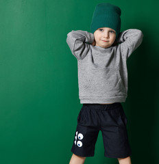 Cool kid boy in blue shorts, gray hoody and knitted hat is posing with his hands behind her head