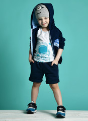 Cool kid boy in blue sweater with his hood on his head stands with his hands in pockets of his shorts and winks