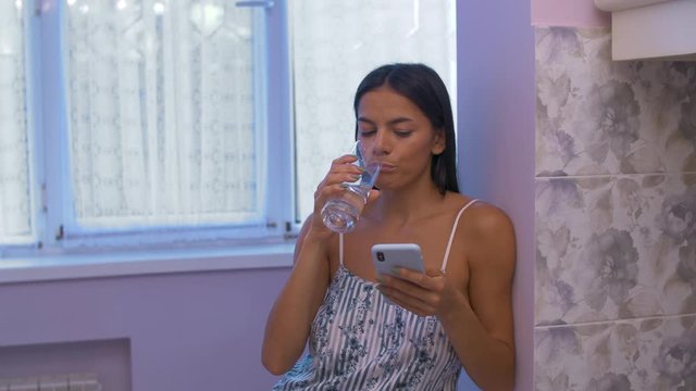 a girl in home clothes in the kitchen drinks water from a glass and leafs through instagram or chatting through her phone sees something unusual, marvels and spits out air the fountain