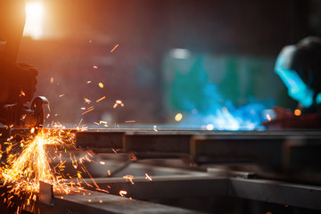 A man grinds metal. In the background a welder welds a part.