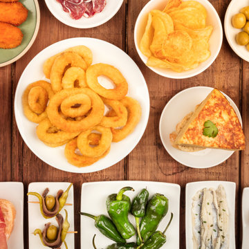 Spanish tapas, a variety of snacks. Squid rings, tortilla, sardines, gildas, padron peppers, square overhead shot on a dark rustic wooden background,