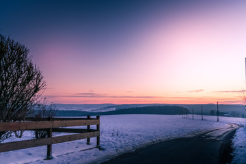 Fototapeta na wymiar Colorful purple and orange sunset over a snowy landscape with rustic wooden fence and winding road over rolling hills