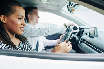 African young woman reading a message on mobile phone and smiling while young man driving a car