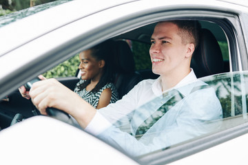 Young smiling man holding the wheel and driving a car with female passenger sitting near by him