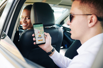 Young man wearing sunglasses holding mobile phone with car sharing app while sitting on back seat...