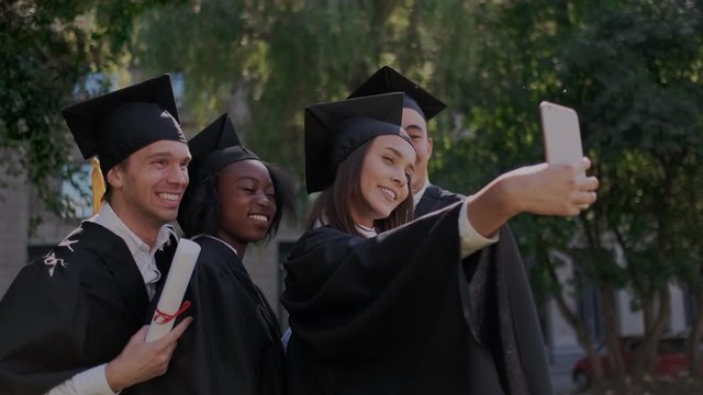 Party of multiethnic graduate students, while making selfie and taking pictures on smartphone. Funny and happy faces, pose on camera. Makes memory. Mantle, academic hats, diploma.