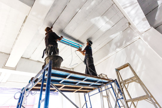 Two technicians standing steel scaffolding is installing fluorescent bulbs on ceiling of building. Home improvement concept.