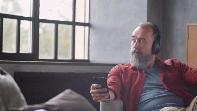Tracking right of old Caucasian man sitting on couch in relaxed pose, listening to music through headphones while holding his smartphone, looking out of window and then looking at camera
