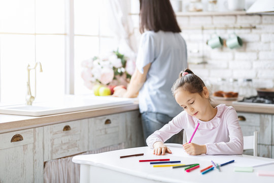 Cute girl drawing at kitchen table while her mother cooking