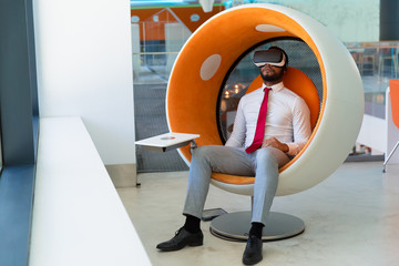 Peaceful businessman in VR headset enjoying virtual video. Man in office clothes and virtual reality goggles sitting in interactive chair and relaxing. Virtual content watching concept