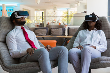 Diverse colleagues in VR glasses relaxing on office lounge. Business man and woman wearing virtual reality headset, sitting in motion chairs. Modern office concept