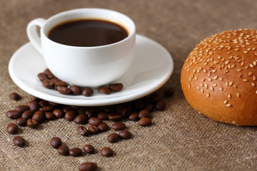 White coffee mug on saucer, bun with sesame seed and scattered coffee beans on burlap tablecloth.