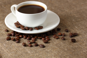 White coffee mug on saucer and scattered coffee beans on burlap tablecloth.