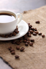 White coffee mug on saucer and scattered coffee beans on burlap tablecloth top view.