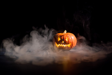 Halloween, orange pumpkin with a scary luminous face on a dark background. Gray thick smoke comes...