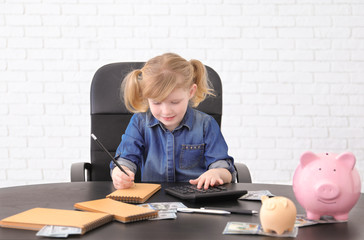 Cute little girl with money and piggy banks at table indoors