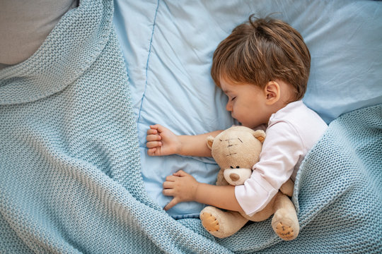 Cute little boy sleeping in bed at home. Sleeping baby in bed, holding a teddy bear. Sweet dreams. Little baby boy sleeping while lying on couch at home. Boy sleeping on bed with teddy bear