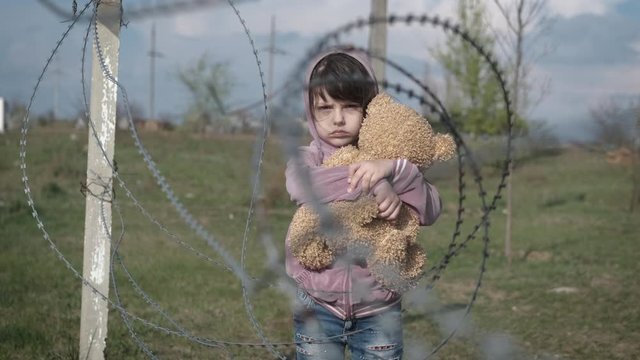 Children's longing. Sad little girl with a teddy bear on the background of barbed wire.