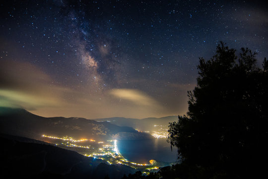 A Starry Sky Hangs Over A Brightly Lit City And Waterway.