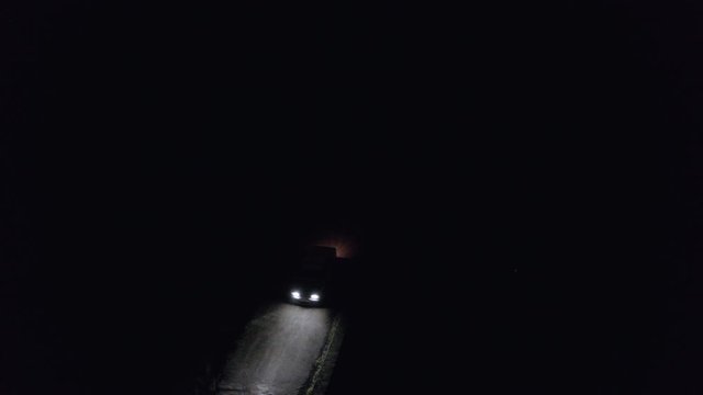 Car moving on countryside road in dark night drone top view. Car headlights illuminating night road in darkness. Night car motion aerial view