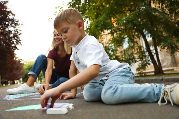 Fototapeta premium Nanny with cute little boy drawing house with chalks on asphalt