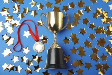 Gold trophy cup, medal and confetti on blue background, flat lay