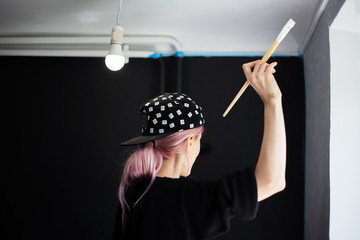 Back view of young girl with pink hair, painting wall with brush and white color, refresh of apartment. Wearing black cap and sweater.