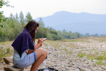A girl sits on the shore of a mountain river and looks into a smartphone. Nearby travel supplies.