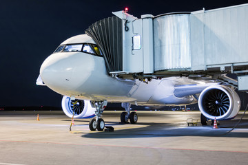 A white passenger aircraft stands at the boarding bridge connected to an external power supply on an airport night apron