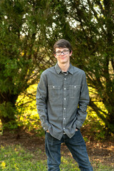 smiling Teenage boy with glasses standing outside in front of a tree wearing a gray button up long sleeved shirt and denim jeans
