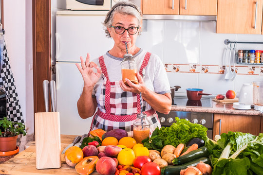 A senior woman with grey hair enjoying the juice fruit just made. Bright light frow the window. Wooden table with a large group of colorful fruits and vegetables. Healthy eating