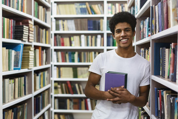 Cheerful african american guy staying between bookshelves, holding book