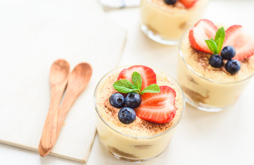 homemade, exquisite dessert tiramisu in glasses decorated with strawberry, blueberry, mint on white wooden table, top view.