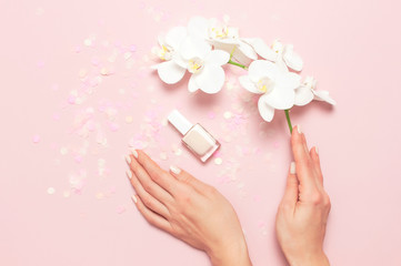 Woman's hands with gentle manicure hold branch of White Phalaenopsis orchid flowers on pastel pink background with festive confetti top view flat lay. Tropical flower. Holiday Women's Day Flower Card