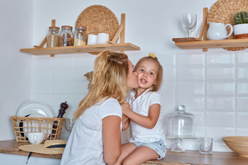 Obraz premium Little girl with mom smile and laugh. A girl sits on the working surface of the kitchen. A cheerful and mischievous daughter with her mother spends time in the kitchen.