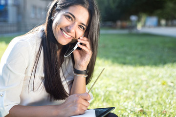 Portrait of a beautiful young indian girl. Student, business woman, sitting on the green grass, talking on the phone and taking notes in a notebook