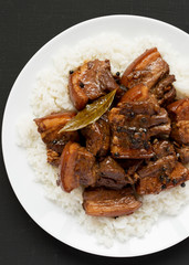 Homemade Filipino Adobo Pork with rice on a white plate on a black surface, top view. Flat lay, overhead, from above. Closeup.