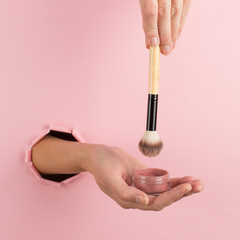 Girl hand holds a brush for makeup and powder from a hole in a pink background. Makeup artist...