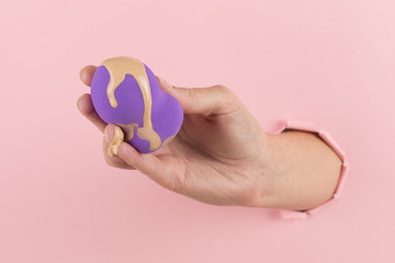 Fototapeta na wymiar Girl hand holds a beauty blender in foundation, concealer from a hole in a pink background. Makeup artist concept, copy space.