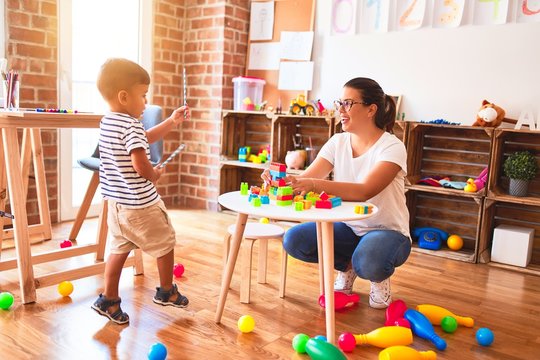 Beautiful teacher and toddler boy playing with construction blocks bulding tower at kindergarten