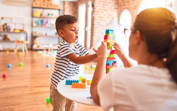 Beautiful teacher and toddler boy playing with construction blocks bulding tower at kindergarten