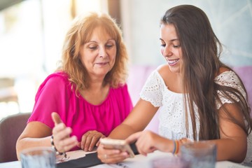 Beautiful mother and daugther sitting at restaurant using smartphone smiling