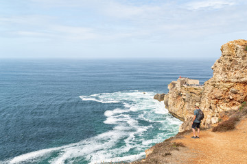Standing man  with a backpack at the edge of a cliff while he is looking at Forte de Sao Miguel in Nazare, Portugal. Wanderlust.